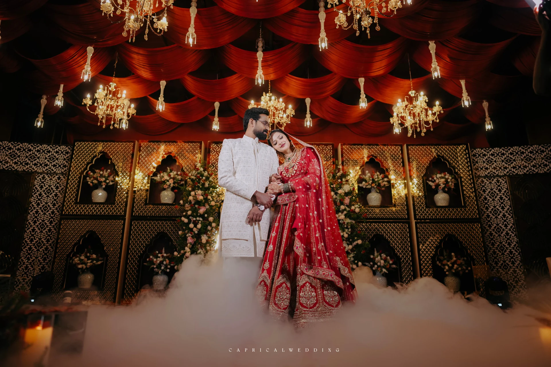 Newlyweds posing beneath red drapery and chandeliers